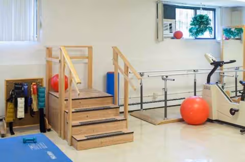 Indoor rehabilitation room with parallel bars, wooden steps, mats, and exercise equipment.