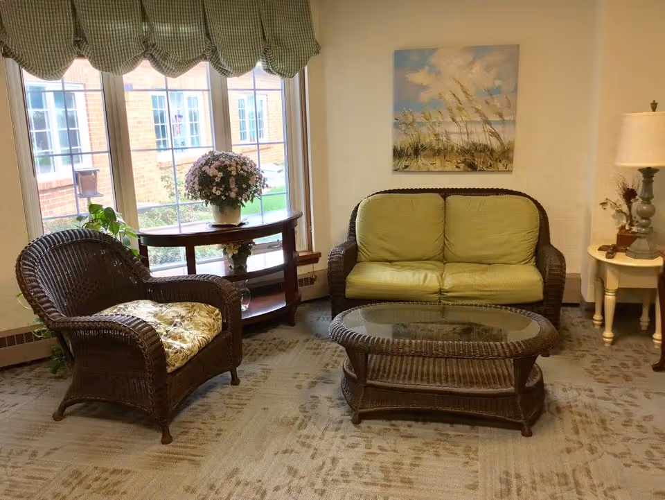 A cozy sitting area with a wicker armchair with floral cushion, a wicker loveseat with green cushions, and a glass-top wicker coffee table. Behind the loveseat is a wall with a painting of a beach scene. To the left, there is a window with green checkered valances and a wooden console table holding a potted plant with pink flowers. To the right, there is a small round side table with a lamp and decorative items.