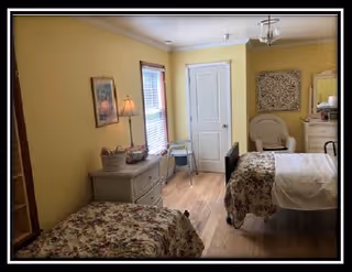A cozy bedroom with two beds covered in floral bedspreads, a small dresser with a lamp and basket, a window with blinds, a white door, and a chair next to a dresser with a mirror. The walls are painted yellow and the floor is wooden.