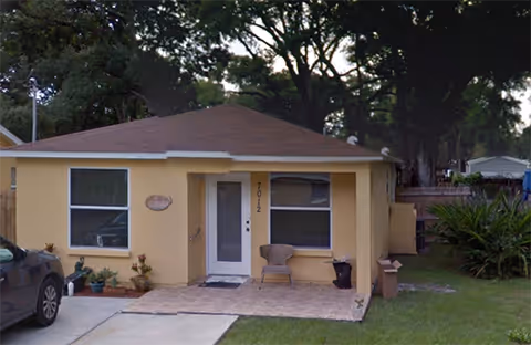 Single-story yellow house with a brown roof, a front door with the number 7012, two windows, a small tiled porch with a chair and a trash bin, a driveway with a parked car, and trees in the background.