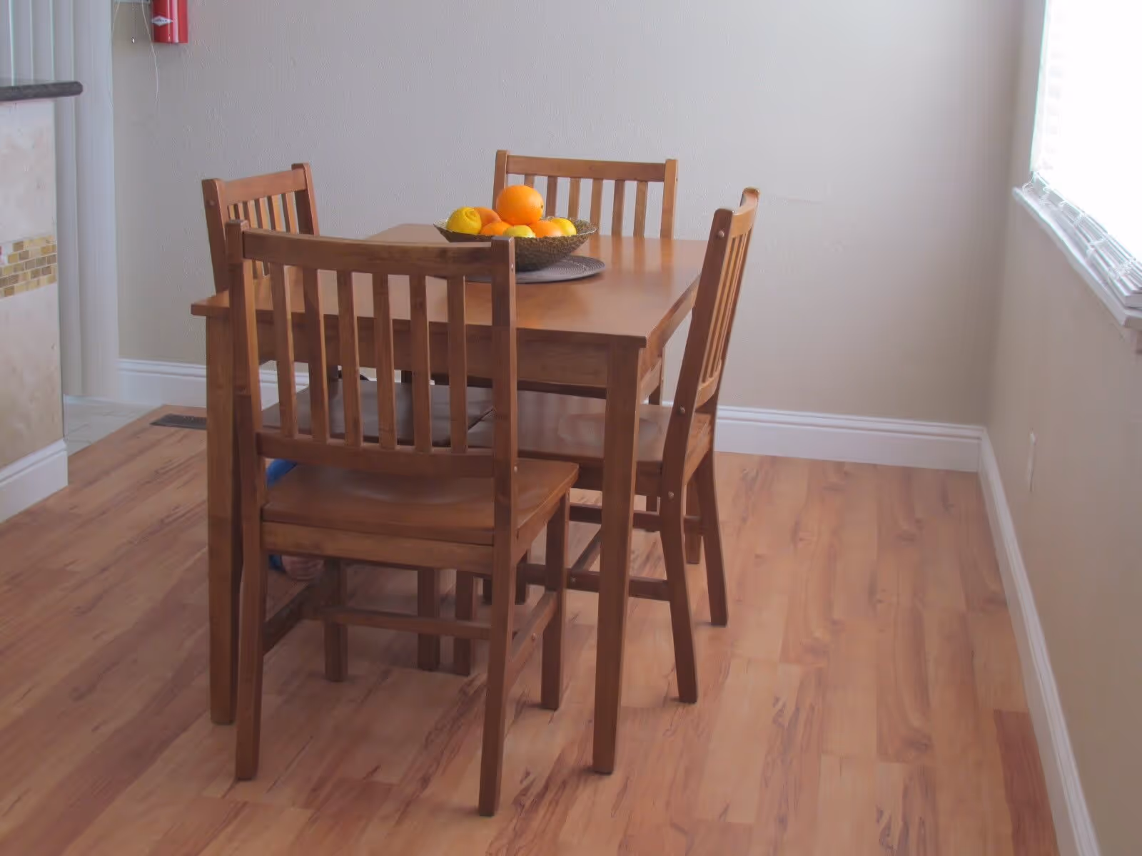 A small dining area with a wooden table and four matching wooden chairs on a light wood floor. A bowl of assorted fruit is placed on the table. The room has beige walls and a window with blinds on the right side.