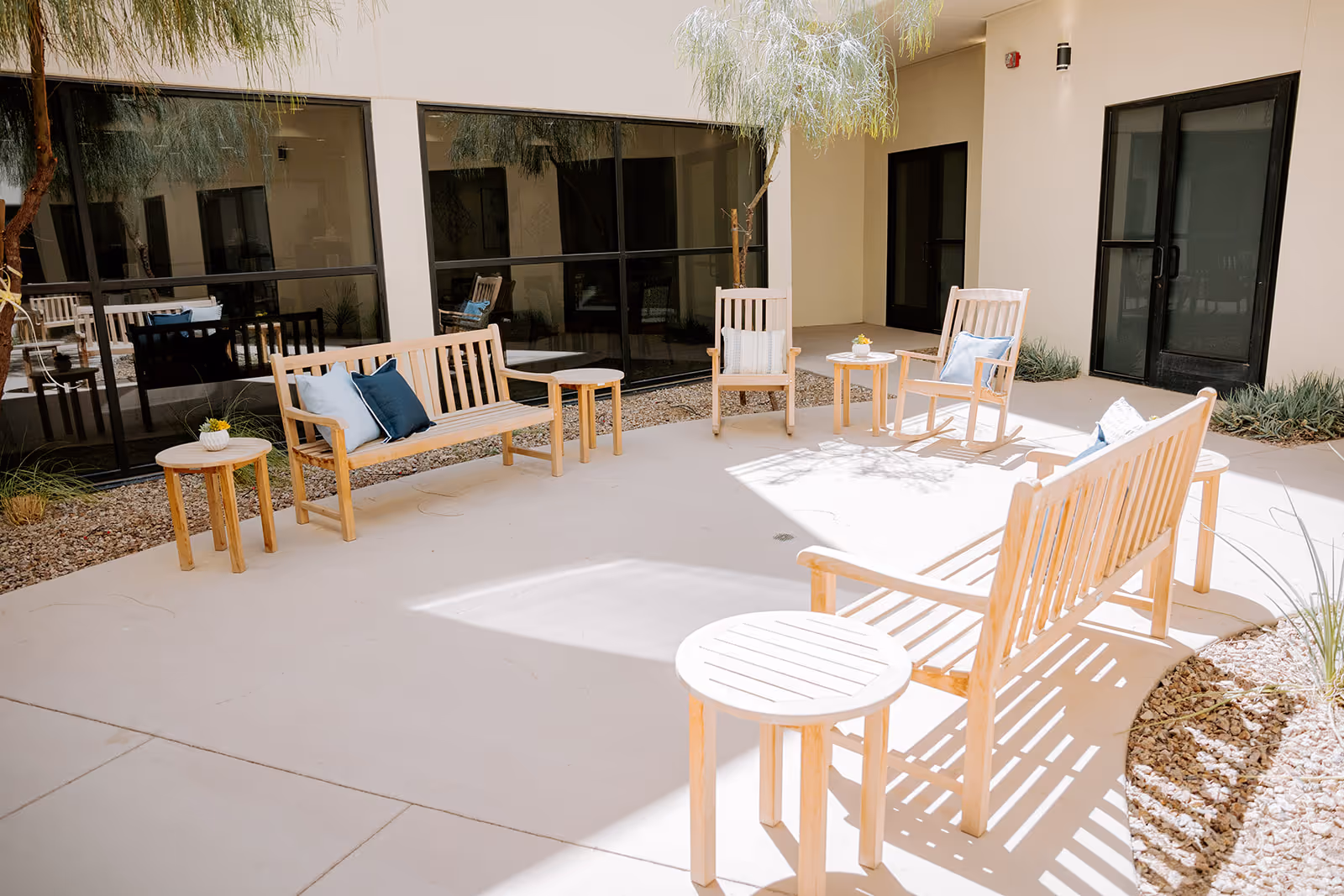 A bright outdoor courtyard area with wooden benches and chairs arranged around small round tables. The seating has cushions in shades of blue and white. The space is surrounded by large windows and doors, with some plants and gravel along the edges.