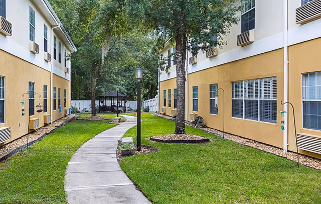 Outdoor walkway between two yellow and white buildings with green grass, trees, bird feeders, and a gazebo in the background.