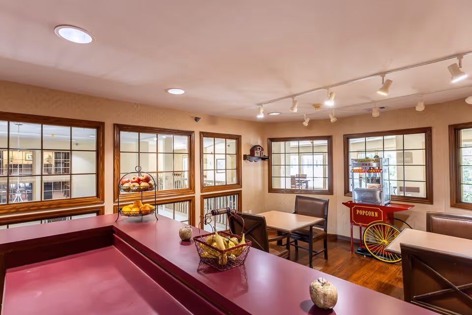 Interior view of a common area in an assisted living facility featuring a maroon counter with baskets of fruit, tables with chairs, large windows with wooden frames, and a popcorn machine on a red cart in the corner.