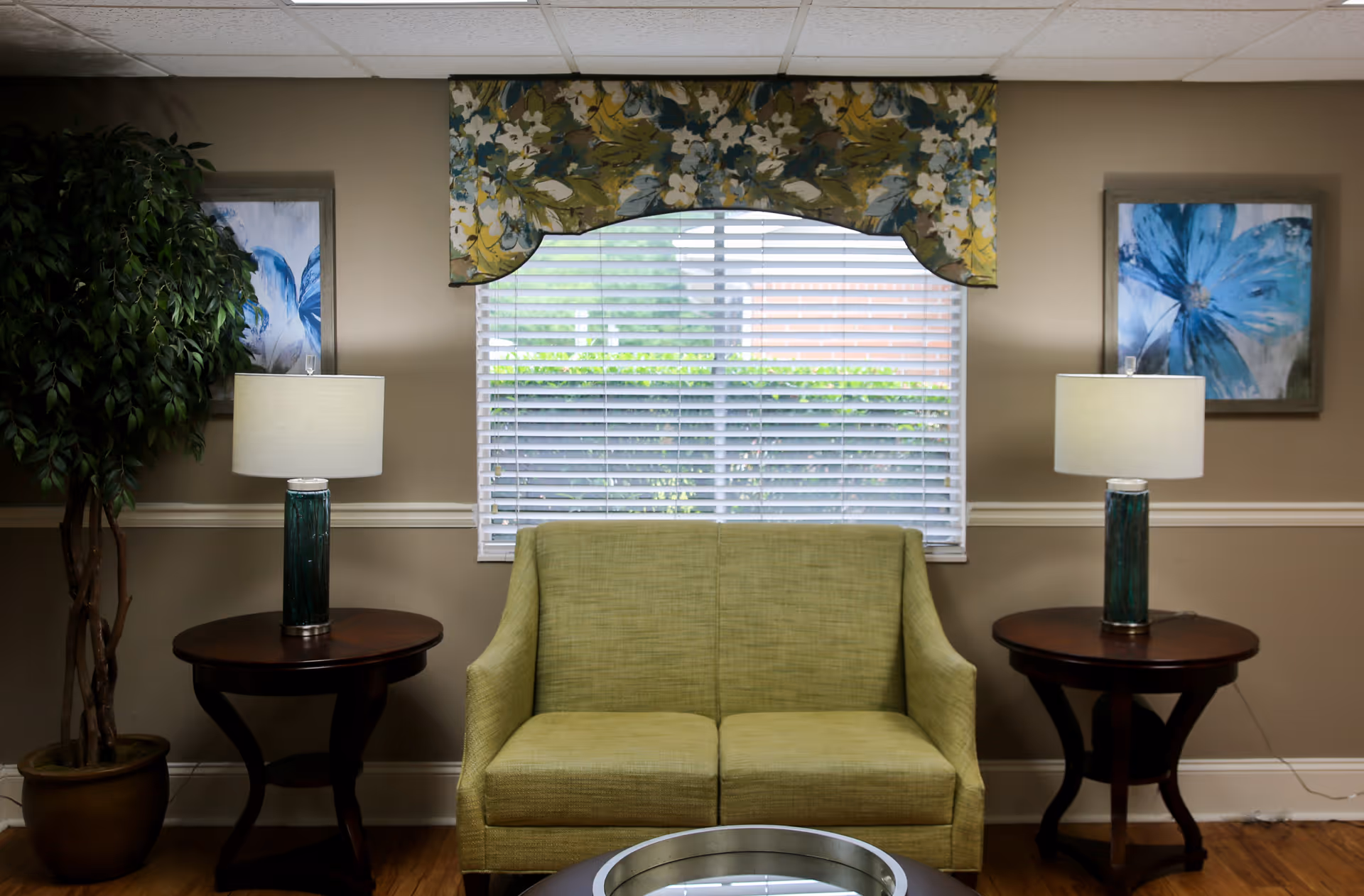 A cozy living room area with a green loveseat centered under a window with white blinds and a floral valance. On either side of the loveseat are dark wooden side tables, each with a green glass lamp with a white lampshade. There are two framed blue floral paintings on the beige walls, and a potted plant is visible on the left side. The floor is wooden.