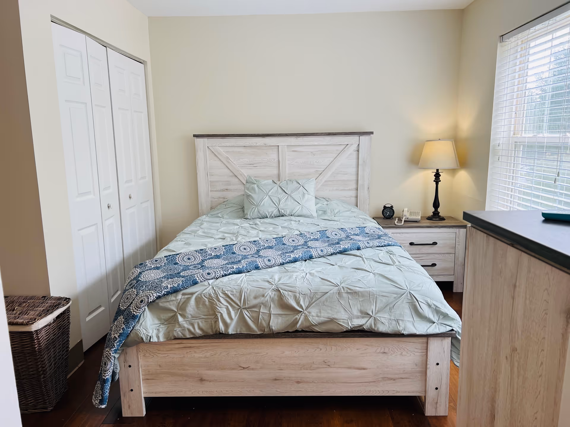A cozy bedroom with a wooden bed frame and a light blue comforter with a matching pillow. A blue patterned throw blanket is draped across the bed. To the right of the bed is a wooden nightstand with a lamp, a clock, and a telephone. There is a window with blinds letting in natural light. On the left side of the room is a white double-door closet and a wicker laundry basket.