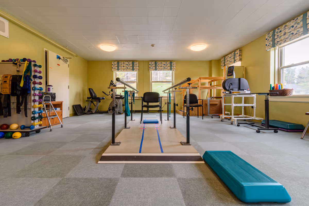 A bright rehabilitation exercise room with parallel bars, therapy equipment, weights, chairs and windows.