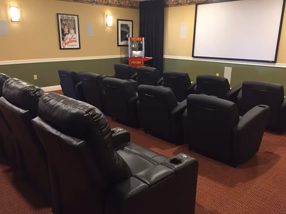 A small movie theater room with two rows of black leather recliner chairs facing a large white projection screen. There is a popcorn machine in the corner and framed movie posters on the walls. The room has beige and green walls with wall sconces providing light.