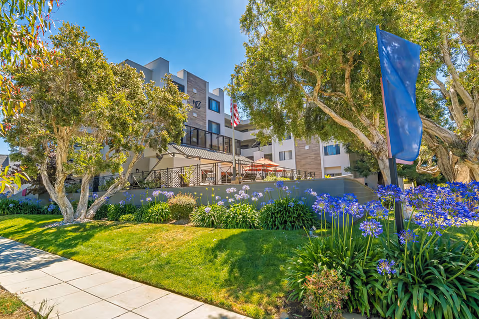 Exterior view of Burlingame Senior Living facility showing a multi-story building with balconies, surrounded by lush green trees, flowering plants, and a well-maintained lawn under a clear blue sky.