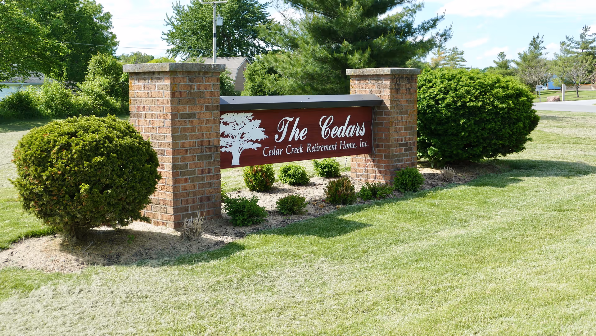 A brick sign reading 'The Cedars Cedar Creek Retirement Home, Inc.' is mounted between two brick pillars on a grassy lawn with shrubs.