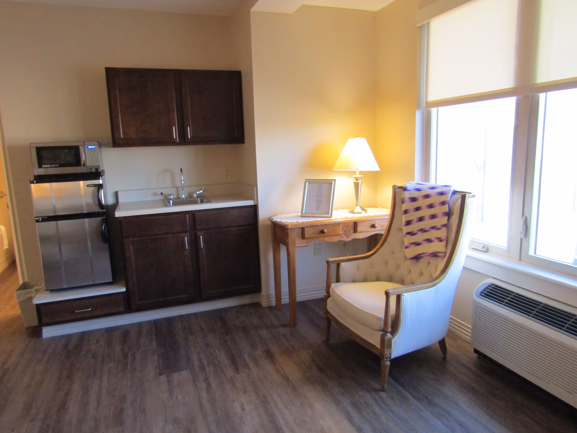 A small kitchenette area with dark wood cabinets, a sink, a mini refrigerator, and a microwave. Next to it is a wooden side table with a lamp and a framed picture. An upholstered armchair with a purple and white blanket draped over the back is positioned near a large window with a white roller shade. Below the window is a wall-mounted air conditioning unit. The floor is covered with wood-look vinyl planks.
