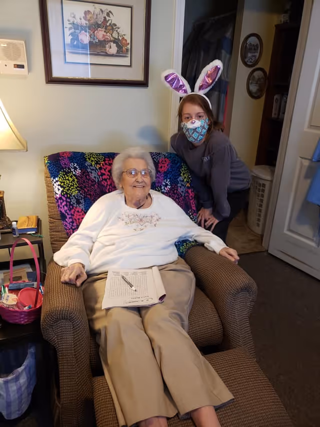 An elderly woman sitting comfortably in a brown armchair with a colorful blanket draped over the back. She is wearing glasses, a white sweater, and beige pants, holding a pen and a puzzle book on her lap. Next to her, a younger person wearing a face mask and bunny ears leans in, smiling. The setting appears to be a cozy living room with a lamp, framed floral artwork on the wall, and various personal items on a side table.