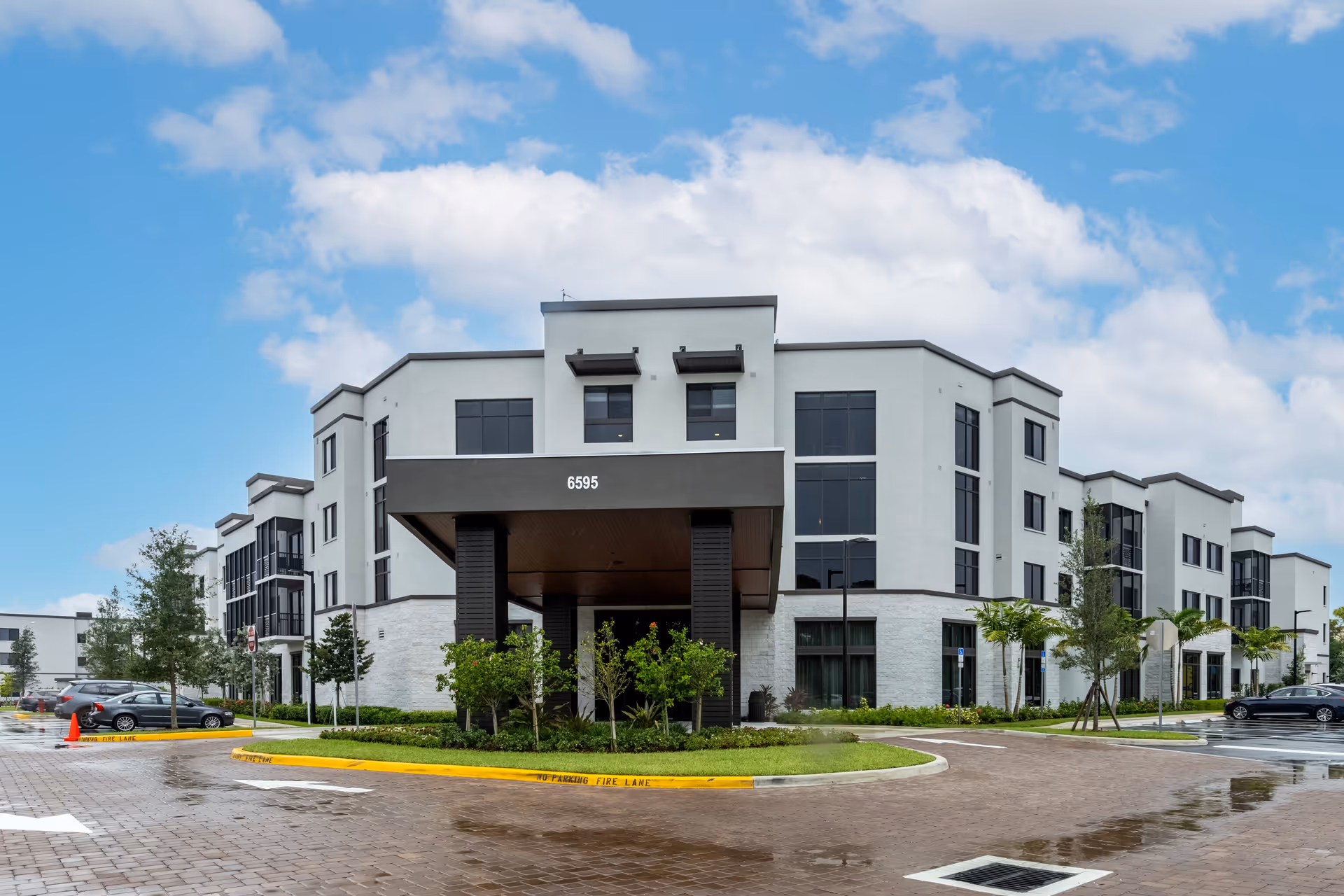 Exterior view of a modern three-story senior living facility building with large windows, a covered entrance with the number 6595 displayed, surrounded by landscaped greenery and a paved driveway with parking spaces.
