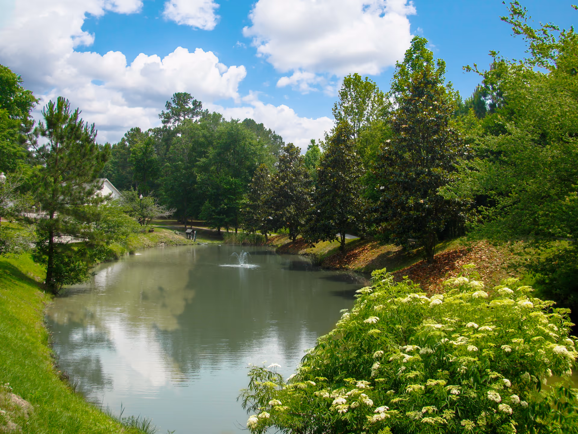 A serene outdoor scene at Cumberland Village featuring a small pond with a fountain in the center, surrounded by lush green trees and bushes under a partly cloudy blue sky.