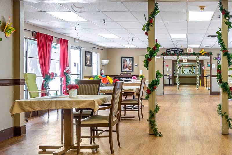 A bright dining area in a senior living facility with tables covered in beige tablecloths and wooden chairs. The room features large windows with red curtains, colorful floral centerpieces on the tables, and decorative green garlands with red flowers wrapped around pillars. The floor is wooden, and there are framed pictures on the walls. The hallway beyond the dining area is decorated with similar garlands and has exit signs visible.