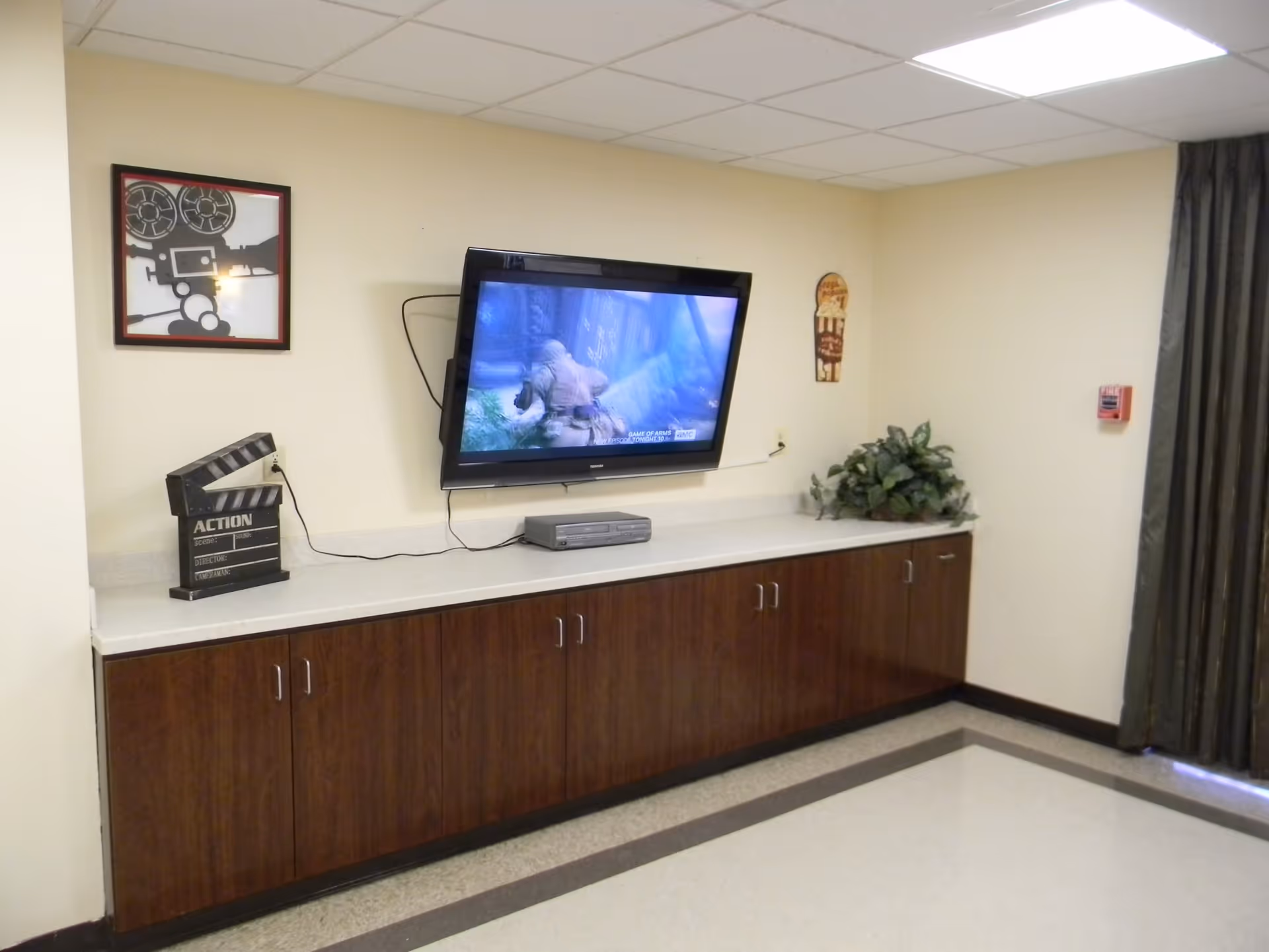 A room with a wall-mounted flat-screen TV displaying a video game scene. Below the TV is a long wooden cabinet with multiple doors. On the cabinet, there is a DVD player, a decorative clapperboard labeled 'ACTION,' and a green plant. The walls are light-colored, and there is a framed picture of a vintage film camera on the wall. A dark curtain covers a window or door on the right side of the image.
