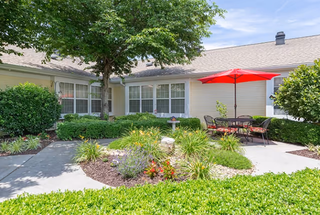 Outdoor courtyard area at TerraBella Morristown featuring a garden with various plants and flowers, a tree providing shade, and a patio with a table and chairs under a red umbrella. The building exterior with windows is visible in the background under a partly cloudy sky.