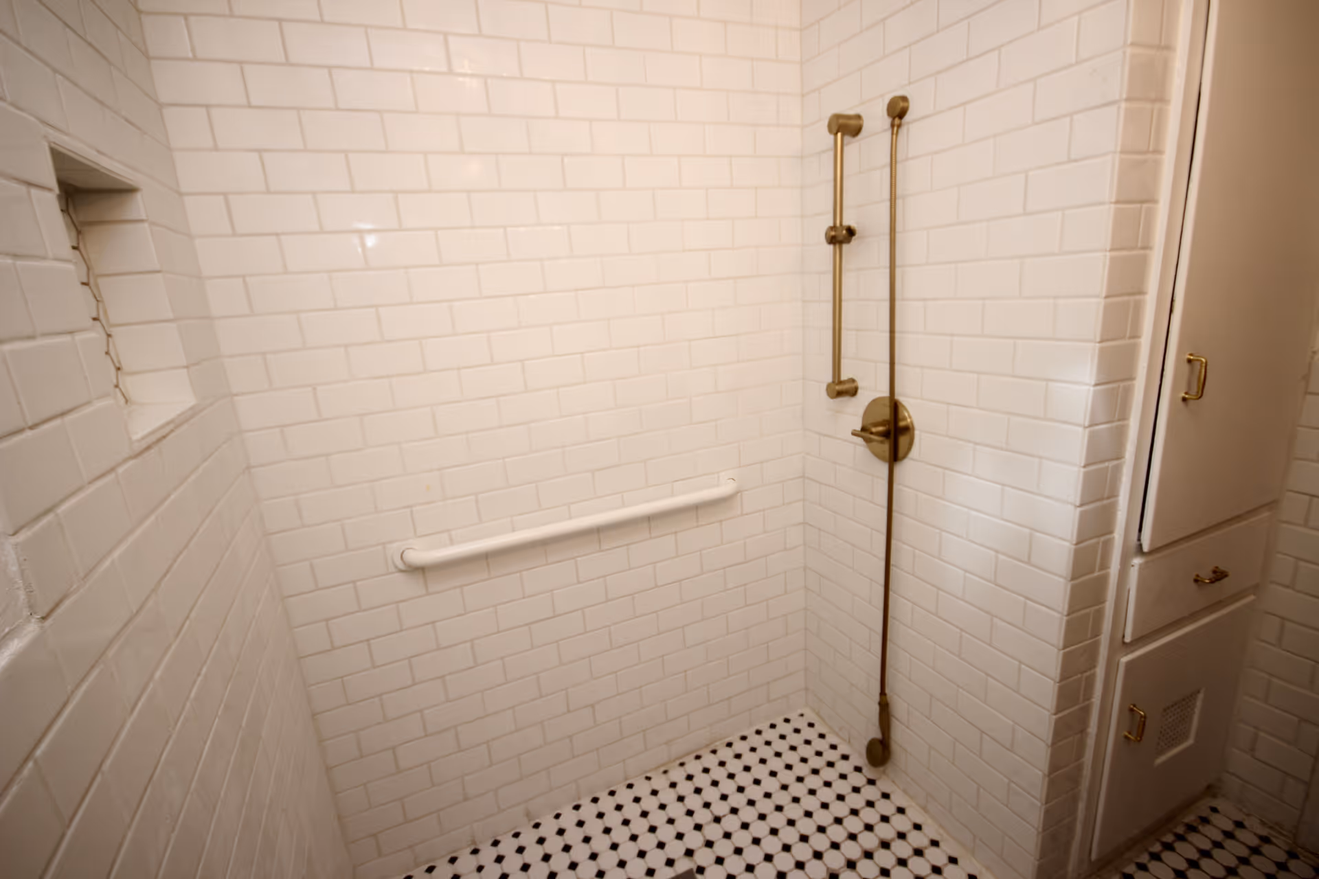 A tiled shower area with white subway tiles on the walls and black and white hexagonal tiles on the floor. The shower features a brass showerhead and handle, a white grab bar on the wall, and built-in shelving. There is a white cabinet with brass handles next to the shower.