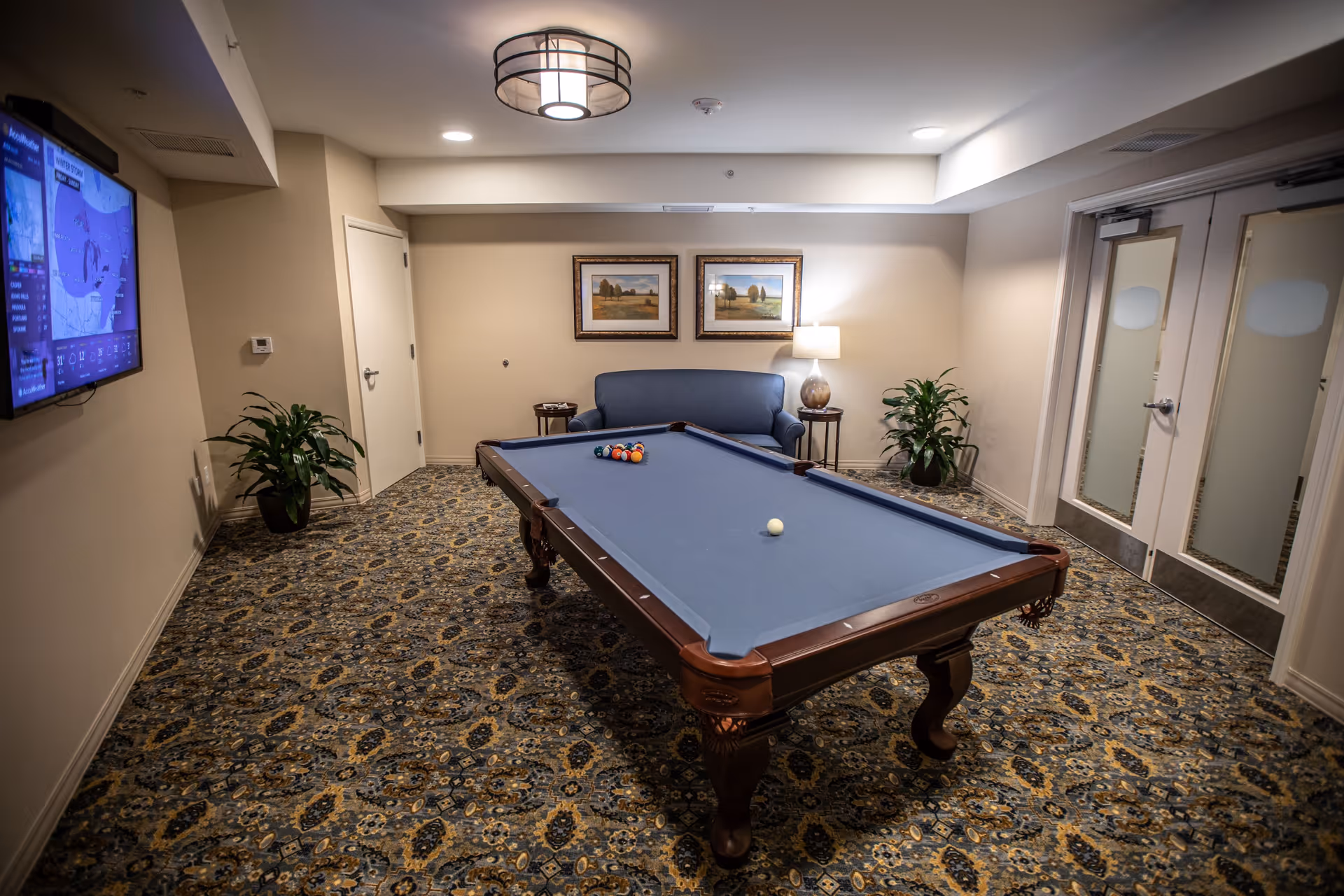 A recreation room with a blue felt pool table in the center, a blue couch against the back wall with two framed landscape paintings above it, a table lamp on a side table to the right of the couch, two potted plants on either side of the couch, a patterned carpet, a wall-mounted TV on the left showing a weather forecast, and double doors with frosted glass on the right.