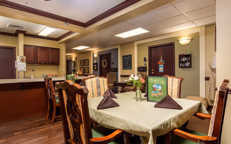 Indoor dining area with a set table, floral centerpiece, upholstered chairs, and a service counter in a senior living facility.