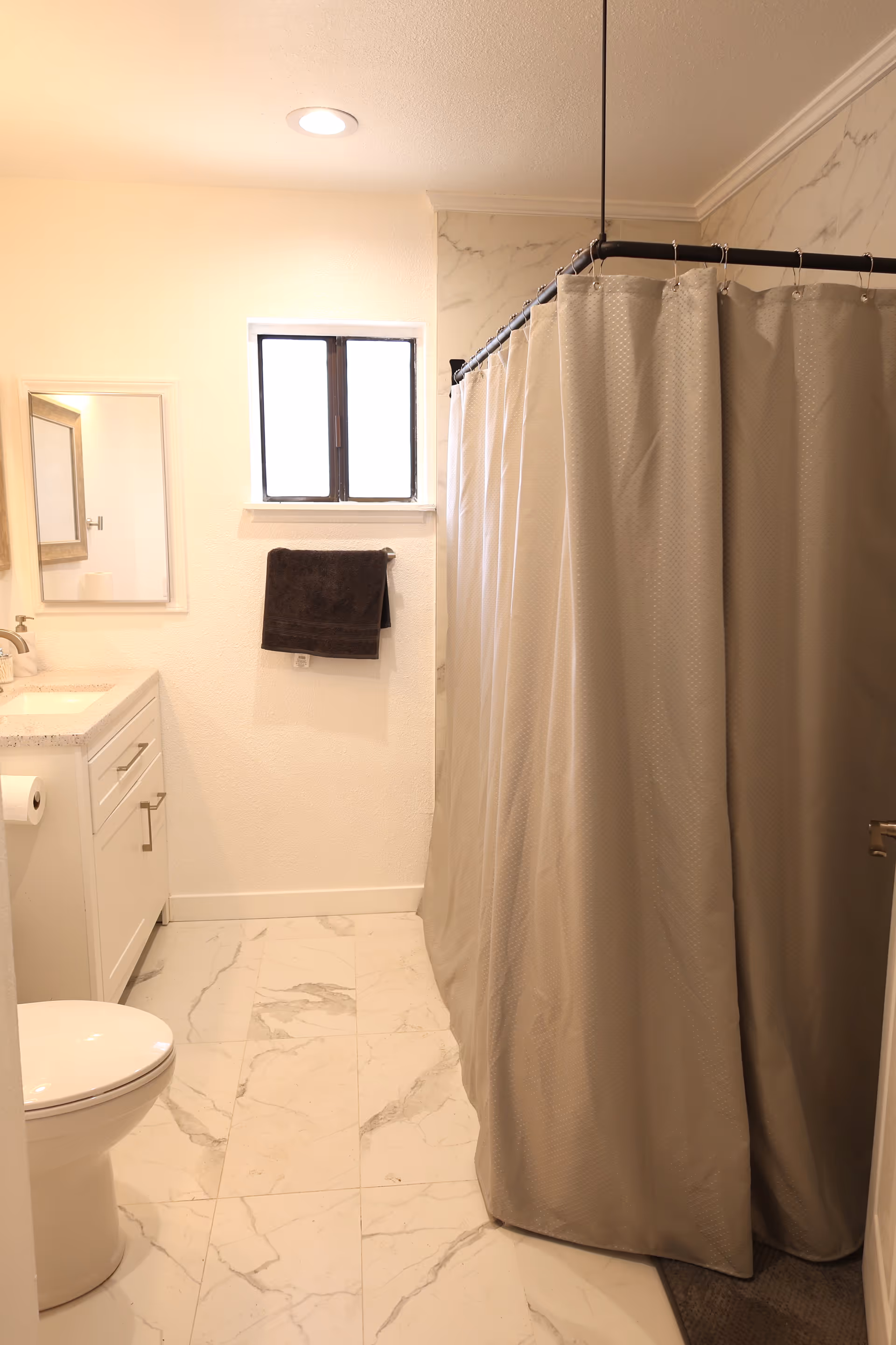 A clean bathroom featuring a white toilet, a white vanity with a sink and mirror, a small window above a towel rack holding a dark towel, and a shower area enclosed by a beige shower curtain. The floor and part of the walls have a light marble pattern.
