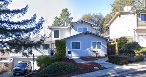 Blue multi-unit residential building with sloped roof, shrubs, and a car parked nearby.