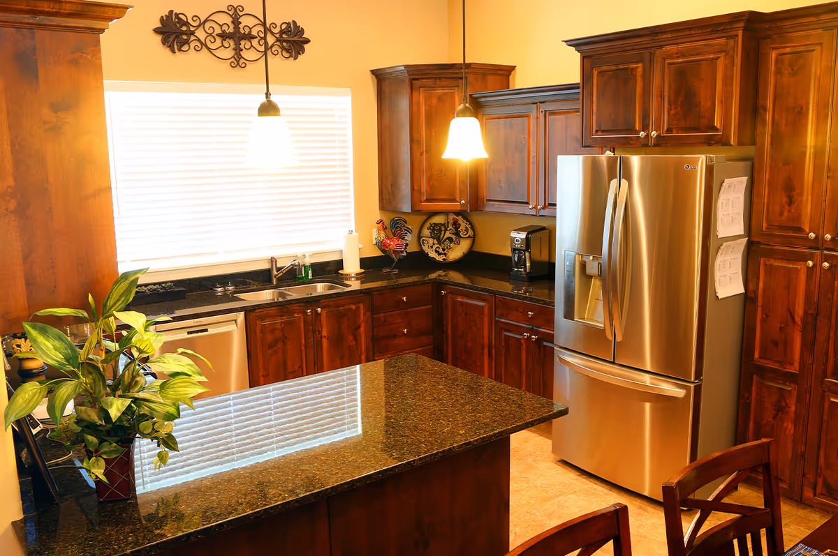 A warm kitchen interior featuring dark wooden cabinets, a stainless steel refrigerator with papers attached, a double sink under a window with blinds, a coffee maker on the counter, and a granite countertop island with a potted plant. Two pendant lights hang from the ceiling illuminating the space.