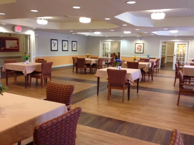 A spacious dining room with multiple tables covered in light pink tablecloths, each surrounded by woven chairs. The room has wooden flooring with a darker wood pattern in the center, soft overhead lighting, framed floral artwork on the walls, and small flower arrangements on each table.