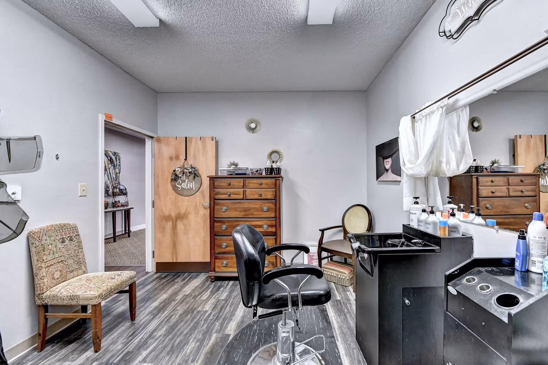 Interior view of a salon room in a senior living facility featuring a black salon chair in front of a black sink and counter with various hair care products. There is a wooden dresser against the back wall, two chairs with patterned upholstery, and a door with a sign that reads 'The Salon'. The room has light-colored walls and wood-patterned flooring.