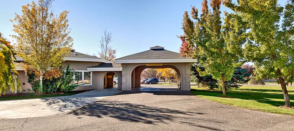 Entrance area of Wellsprings Assisted Living facility with a covered driveway, surrounded by trees with autumn foliage and a clear blue sky.
