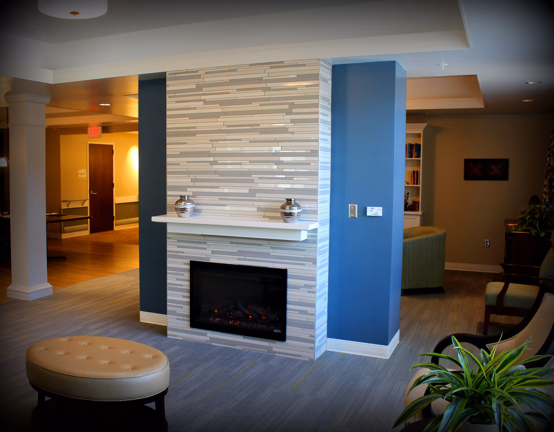 A cozy interior living area featuring a modern electric fireplace with a tiled surround and a white mantel holding two decorative silver vases. The room has blue and beige walls, a beige cushioned ottoman, a green armchair, and a plant in the foreground. There is a hallway with wooden flooring and a door in the background.
