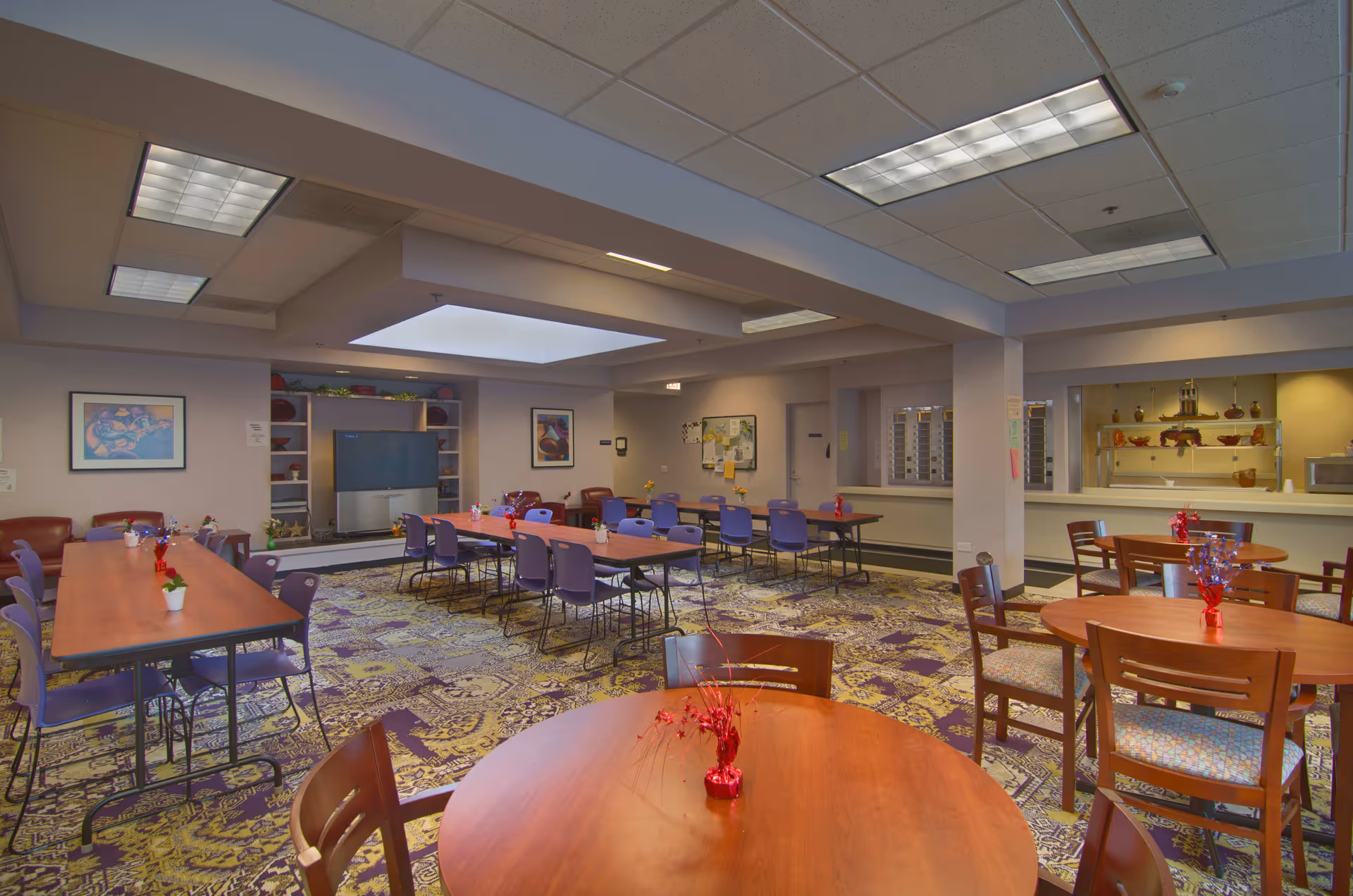 A spacious dining room in a senior living facility with multiple tables and chairs arranged for group seating. The room features a patterned carpet, overhead fluorescent lighting, and a large TV on a stand against the far wall. There are framed artworks on the walls and a counter area that appears to be a serving or reception window.
