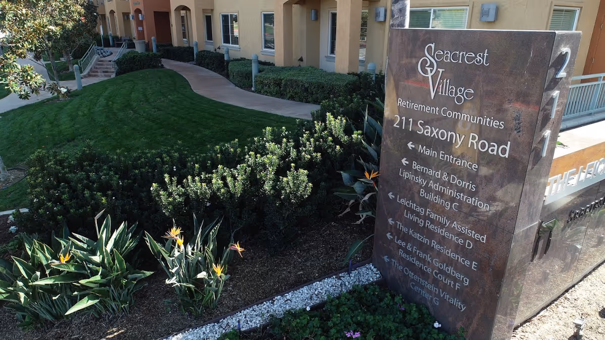 Stone entrance sign for Seacrest Village Retirement Communities at 211 Saxony Road beside landscaped lawn and the building facade.