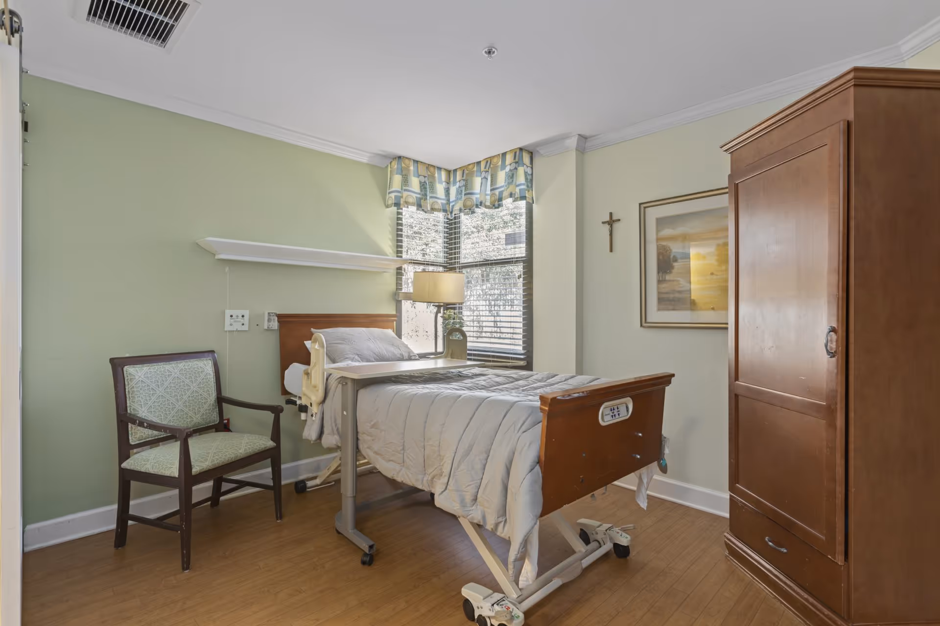 A small, clean nursing facility bedroom with a hospital bed covered in a light gray comforter, a wooden chair with green patterned upholstery, a wooden wardrobe, a lamp on a bedside table, and a window with blinds and a patterned valance. A framed landscape picture and a cross hang on the light green walls.