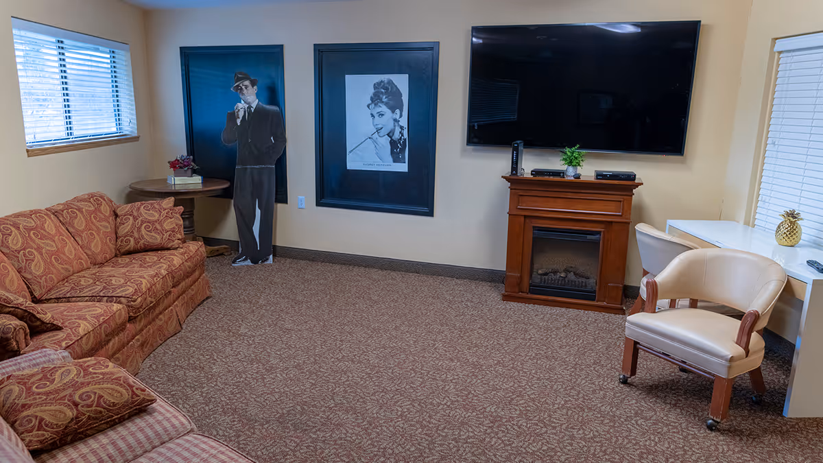 Cozy living room with patterned sofas, an armchair and desk, a large wall-mounted TV above a wooden electric fireplace, and framed portraits on the wall.