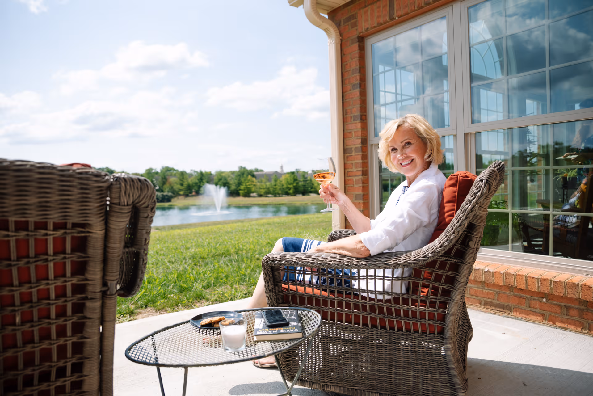 An elderly woman sitting on a wicker chair on a patio outside a brick building, holding a glass of wine and smiling. In front of her is a small round metal table with a book, a smartphone, a candle, and a plate with cookies. In the background, there is a grassy area with a pond and a water fountain under a partly cloudy sky.