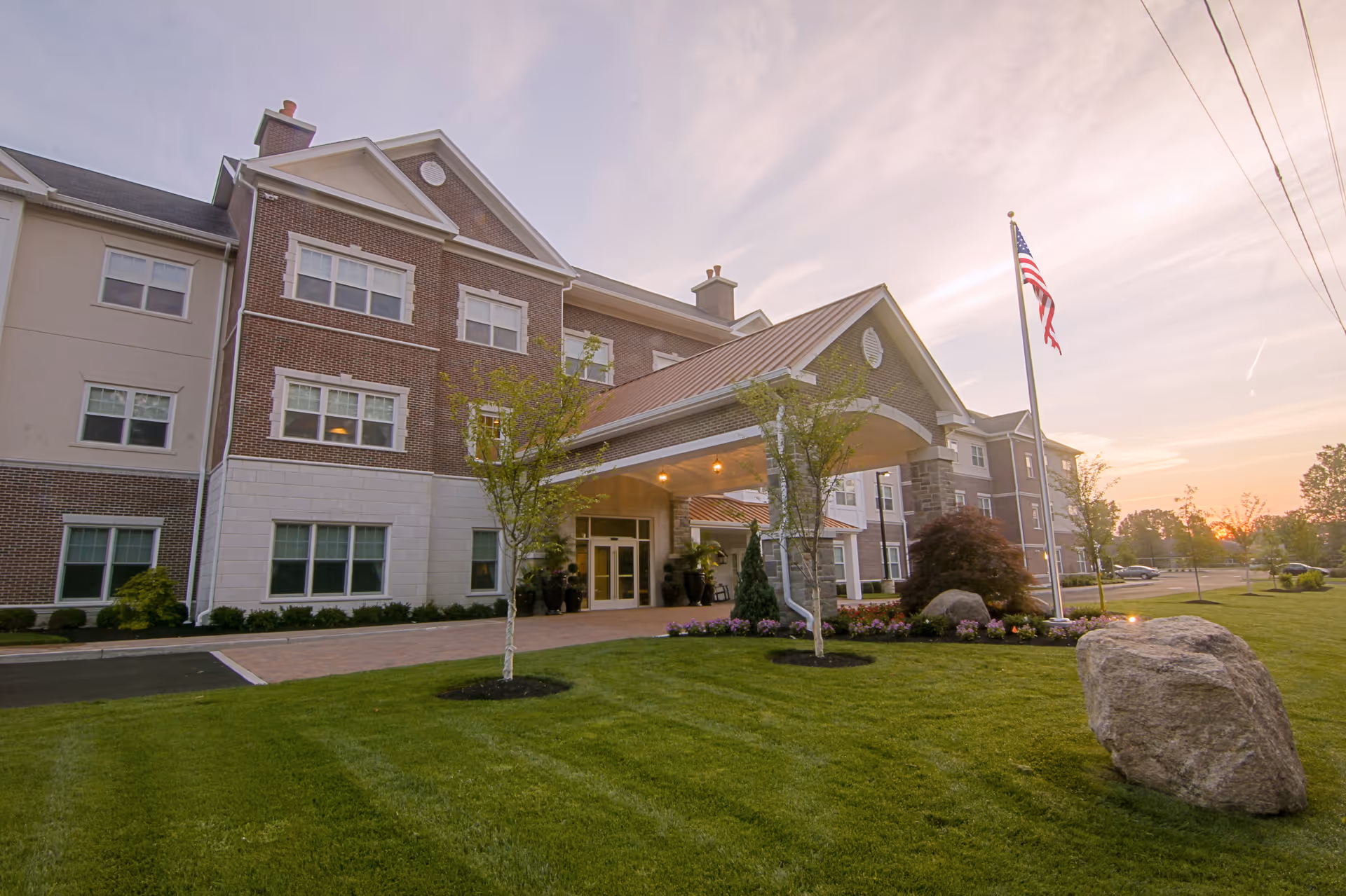 Exterior view of a multi-story senior living facility building with a covered entrance, well-maintained lawn, small trees, flower beds, and an American flag on a flagpole during sunset.