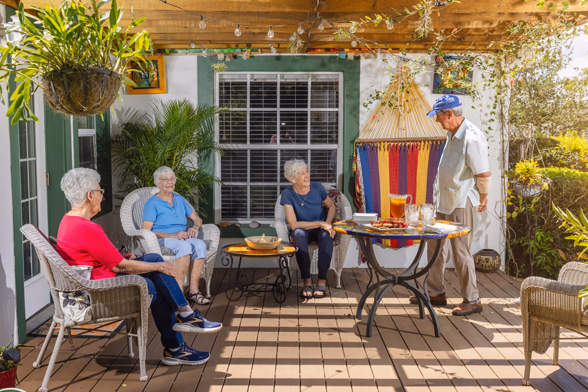 Four seniors sitting and chatting on a shaded outdoor patio with wicker chairs, a round table with drinks and snacks, hanging plants, and a colorful hammock.