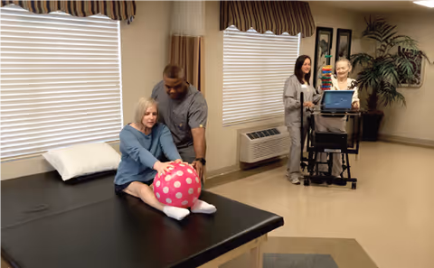 A therapy room where a woman is seated on a black therapy table stretching and holding a large pink ball with white polka dots, assisted by a male therapist. In the background, an elderly woman is using a walker with a tray holding a laptop, accompanied by a female caregiver. The room has beige walls, two windows with blinds and striped valances, framed pictures on the wall, and a large potted plant.