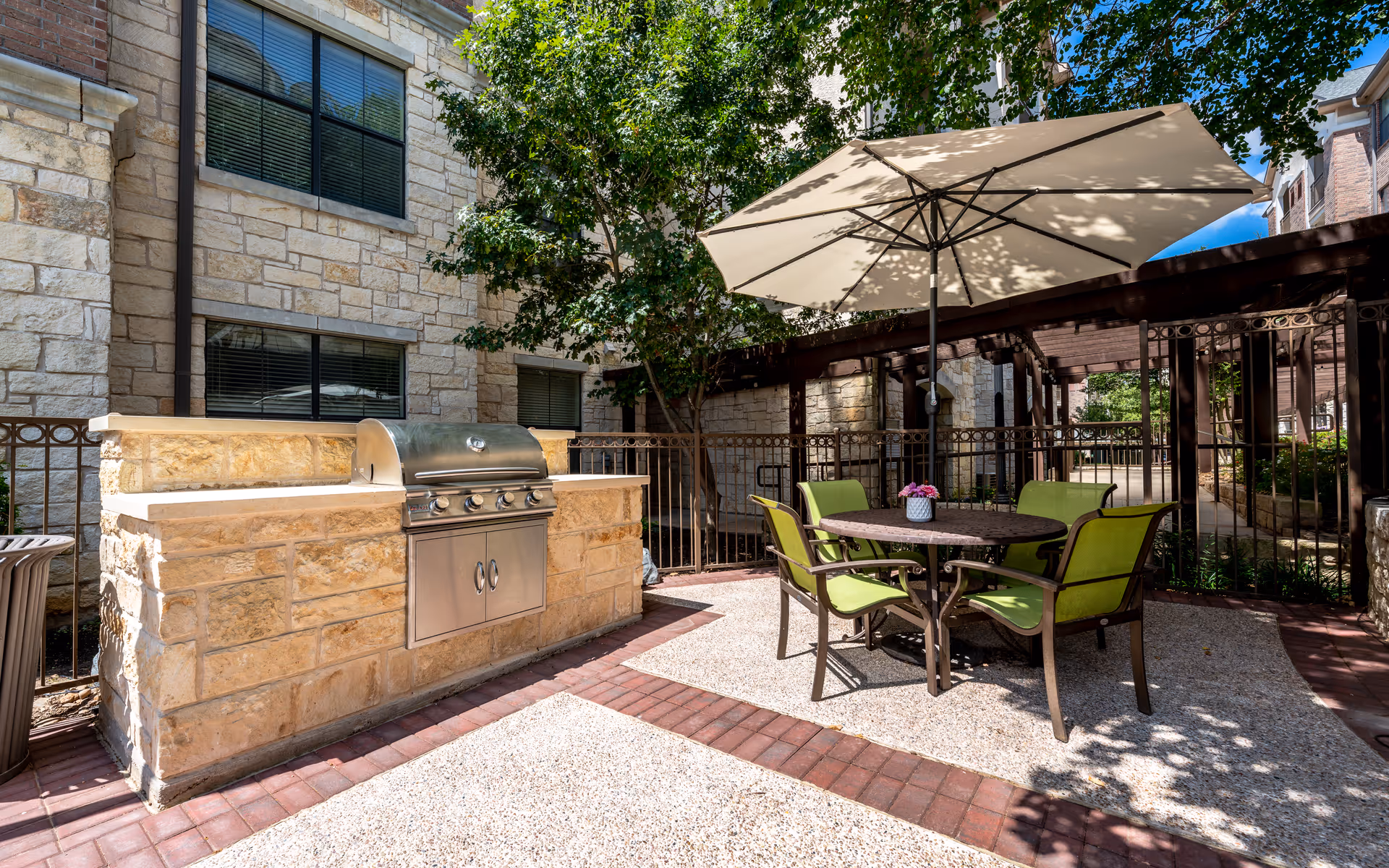Outdoor patio area with a built-in stone grill, a round table with four green chairs, and a large beige umbrella providing shade. The area is surrounded by a metal fence and stone buildings with windows, and there are trees providing additional shade.