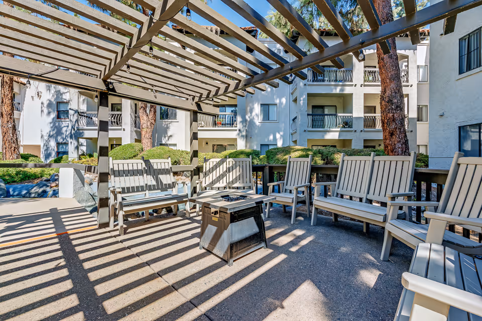 Outdoor seating area at a senior living facility featuring multiple beige wooden chairs and benches arranged around a central fire pit under a pergola with string lights. The background shows white multi-story buildings with balconies and greenery.