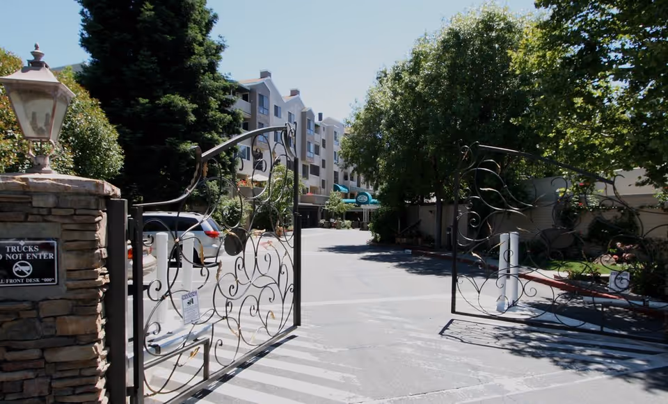 Entrance gate of The Heritage Downtown facility with ornate black metal gates open, a stone pillar with a lamp on the left, and a sign that says 'Trucks Do Not Enter'. In the background, there are multi-story buildings, trees, parked cars, and a clear blue sky.
