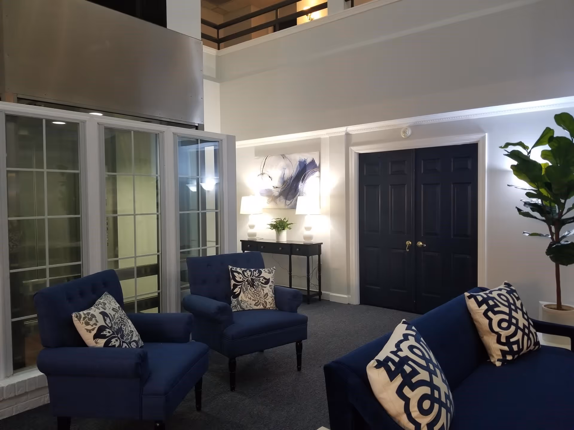 A cozy seating area with two blue armchairs and a blue sofa, each adorned with patterned throw pillows. Behind the chairs is a small black console table with two white lamps and a small plant. Above the table hangs an abstract painting. The room features dark double doors, large windows with white frames, and a tall green potted plant in the corner.