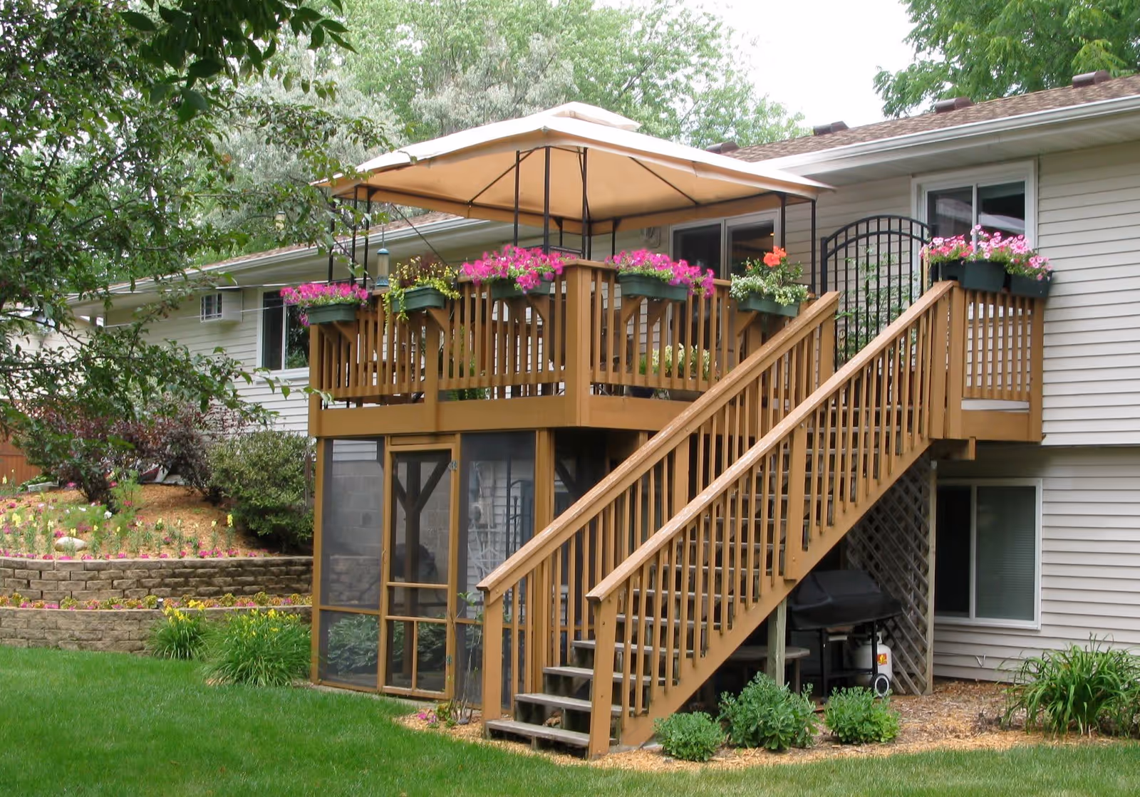 Exterior view of a residential building with a wooden deck and staircase leading up to it. The deck is decorated with flower boxes filled with pink and red flowers and has a beige canopy overhead. Below the deck is a screened-in porch area. The surrounding yard is green with grass, shrubs, and trees.
