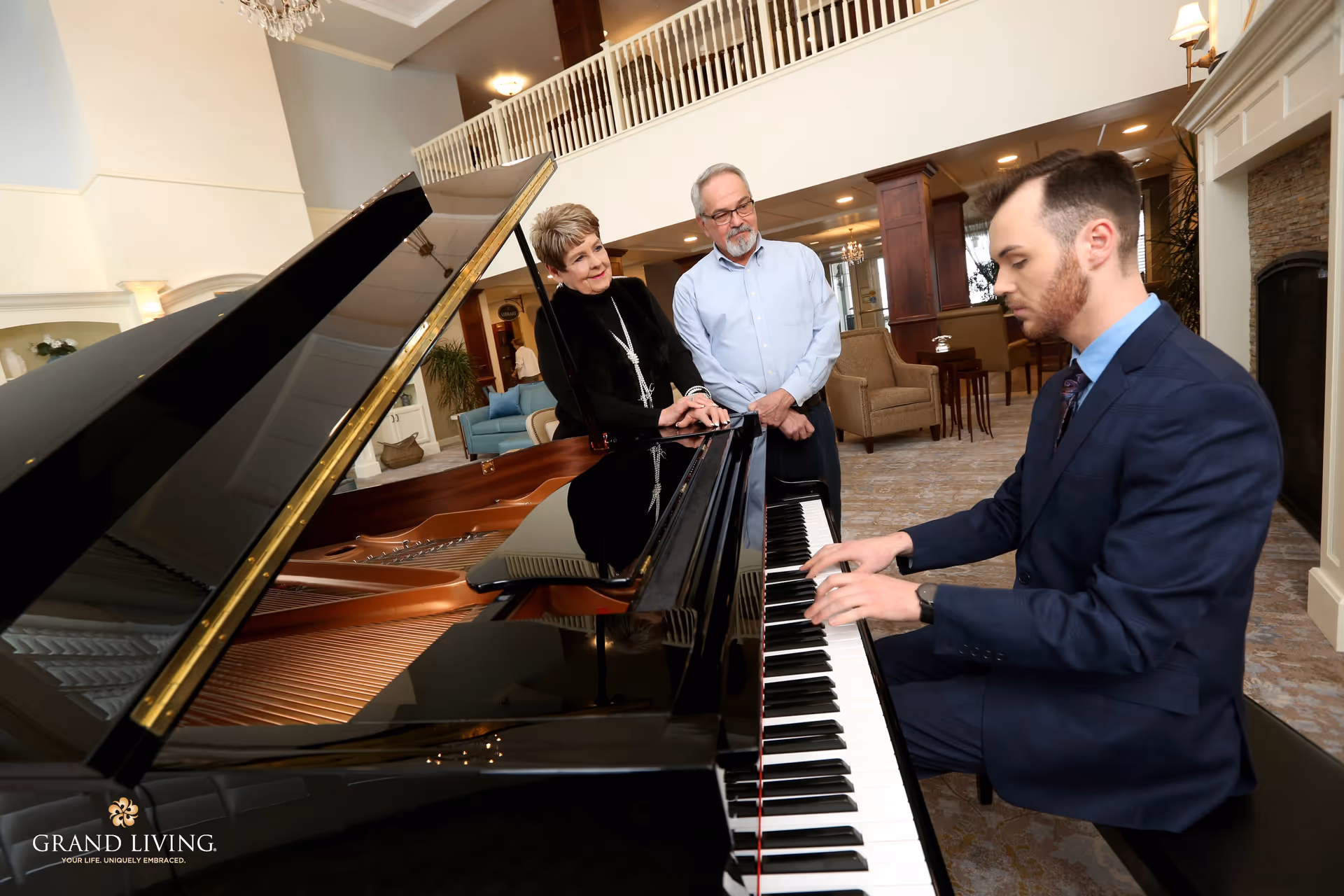 A man in a suit plays a grand piano while an older couple watches in a spacious senior living lounge.