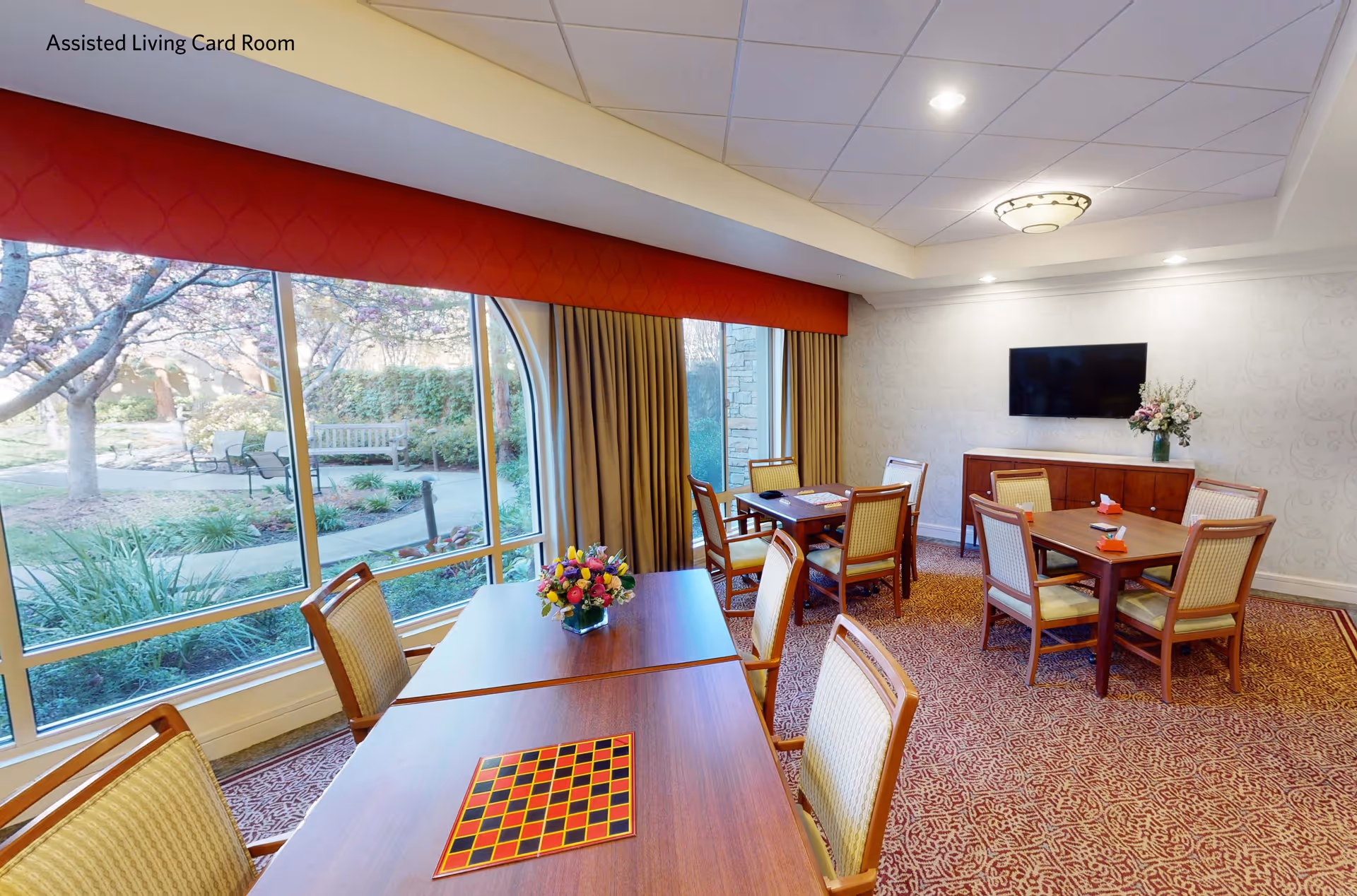 A bright assisted living card room with large windows overlooking a garden. The room contains several wooden tables with chairs arranged around them. One table has a checkerboard on it, and there are flower arrangements on two tables. A flat-screen TV is mounted on the wall above a wooden cabinet with a flower vase. The carpet has a red and beige pattern, and the ceiling has recessed lighting and a decorative light fixture.
