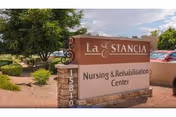 Outdoor view of a sign for La Estancia Nursing & Rehabilitation Center, surrounded by desert landscaping with trees, bushes, and gravel, with a partly cloudy sky in the background.
