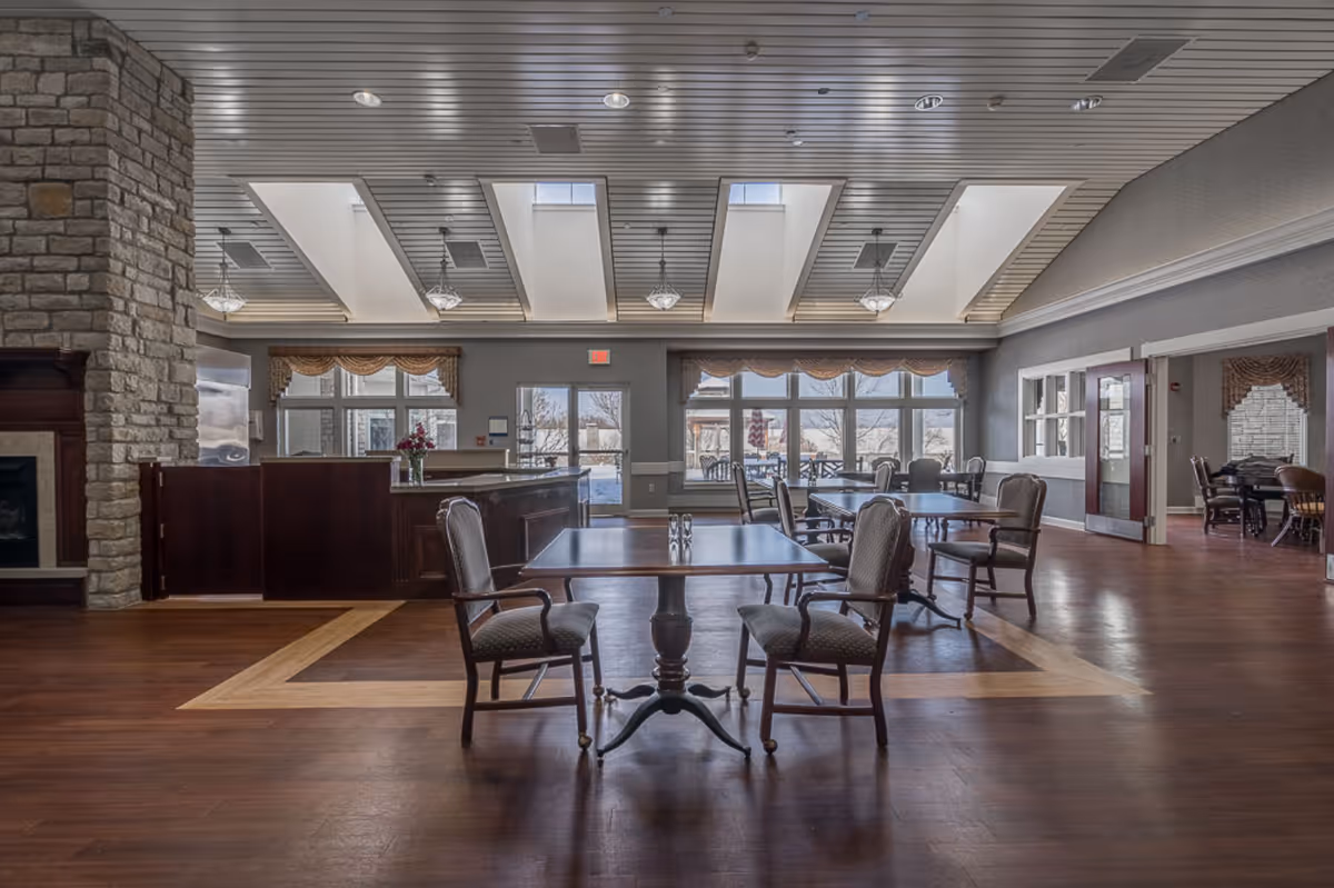 A spacious and well-lit dining area with several wooden tables and cushioned chairs arranged neatly. The room features large windows with valance curtains allowing natural light to enter, a stone fireplace on the left, and a ceiling with multiple skylights and hanging light fixtures. The floor is wooden with a lighter wood inlay design.