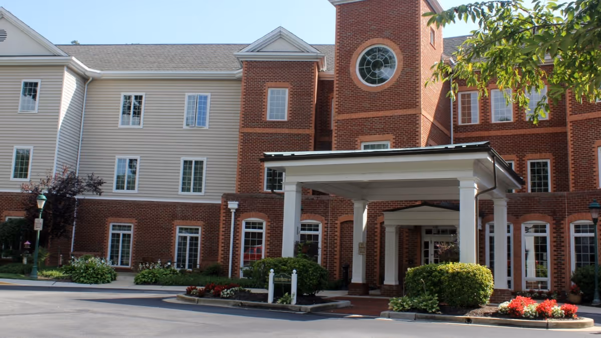 Front entrance of a three-story red brick assisted living building with a covered porte-cochere and landscaped beds.