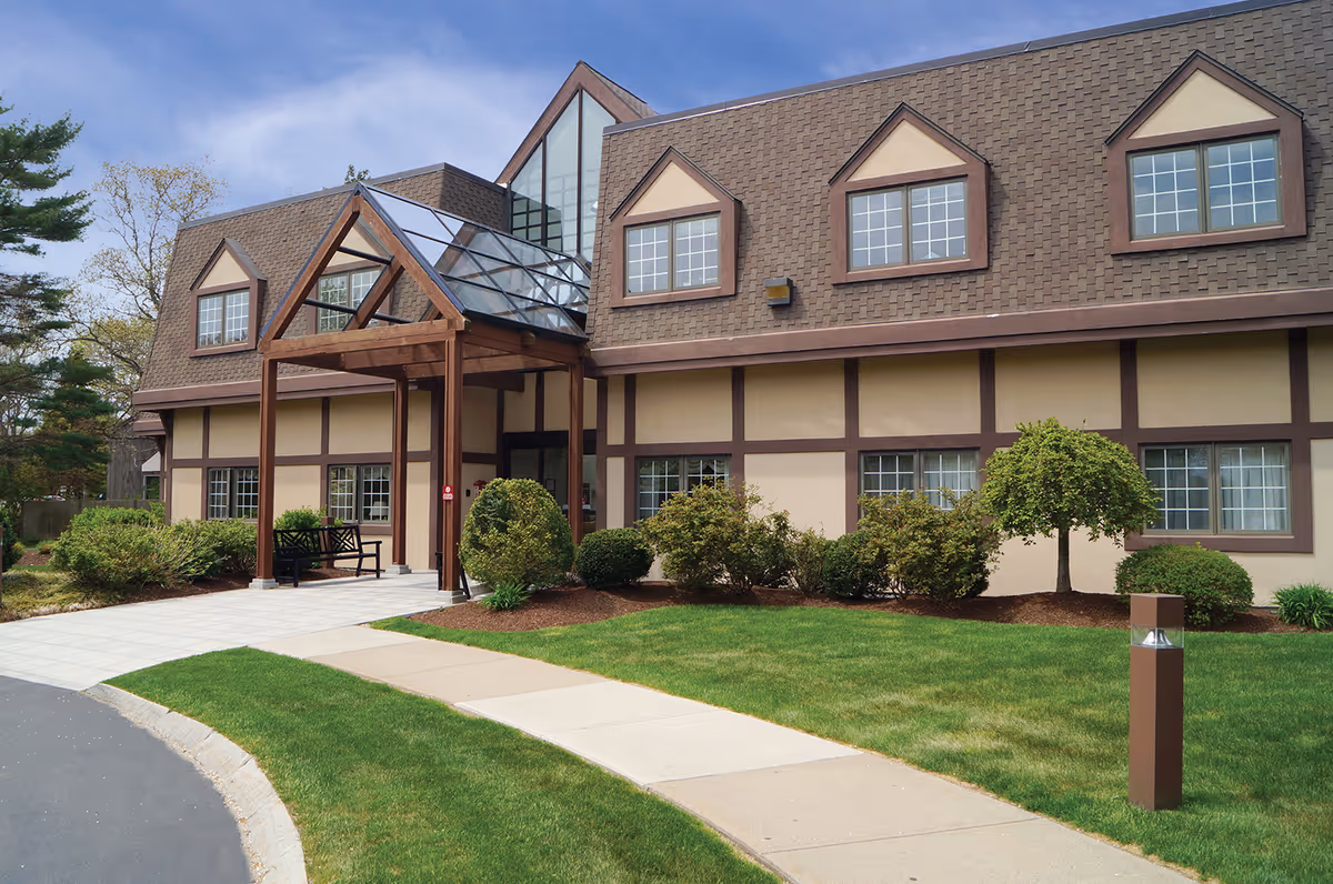 Exterior view of Sippican Health Care Center Inc building with a brown roof, beige walls with brown trim, multiple windows, a glass-covered entrance, and landscaped bushes and grass in front.